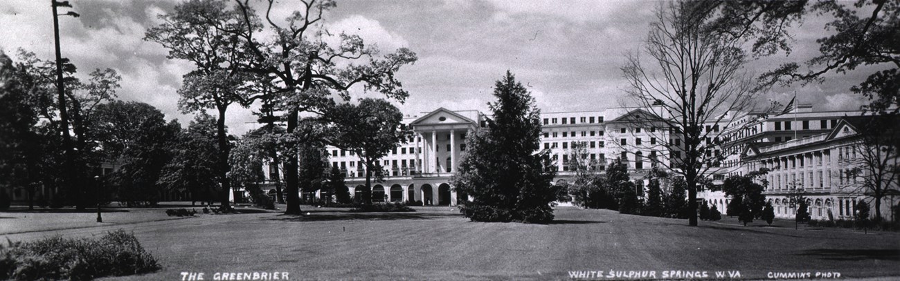 Black and white photo of front lawns and facade of Neoclassical hospital building. Central section has portico on top of columns and is flanked by two multi-story wings. It is surrounded by trees.