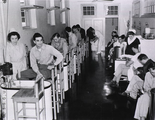 Female nurses stand on either side of an aisle to treat the injured arms and legs of male soldiers. The soldiers are seated on stools next to large metal basins filled with water.