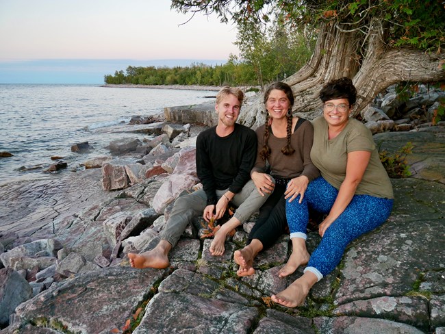 Man and two women in bare feet sit together on a rocky shoreline