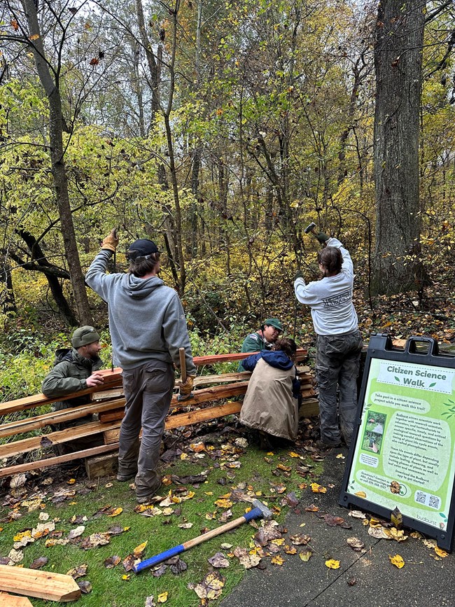 Five individuals help replace a fence. A sign for a citizen science walk is on the right. In the background is the woods.