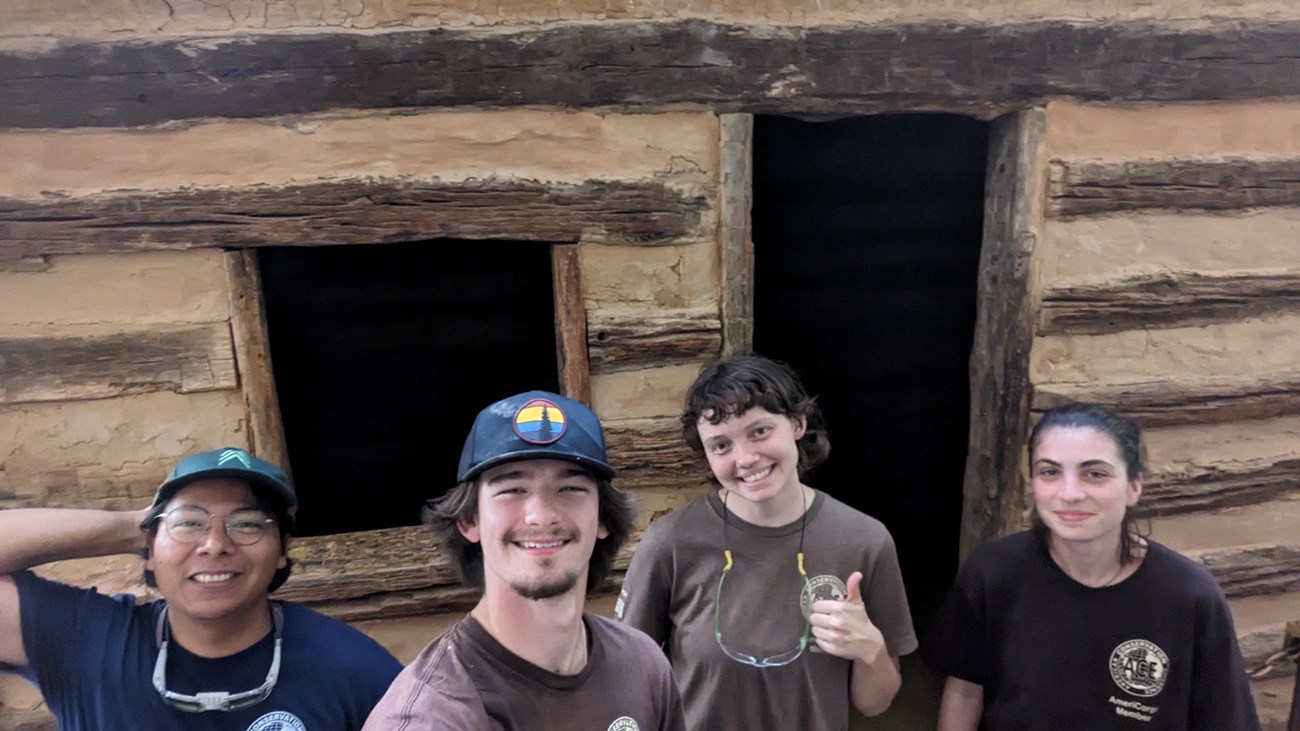 Four young individuals smile in front of a log cabin