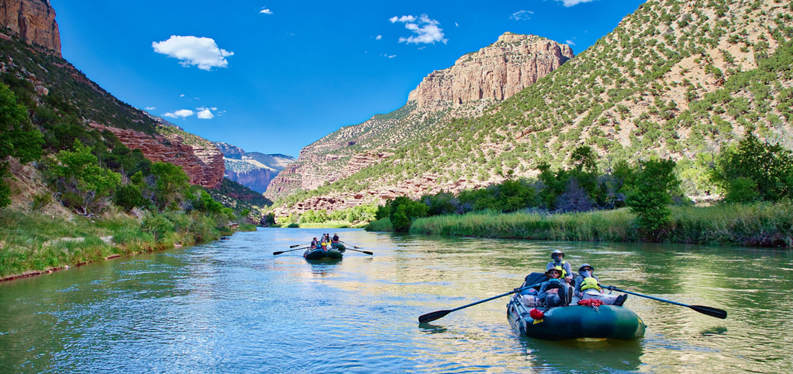 People in rafts float down a calm river surrounded by steep canyon walls and lush greenery under a bright blue sky with scattered clouds.