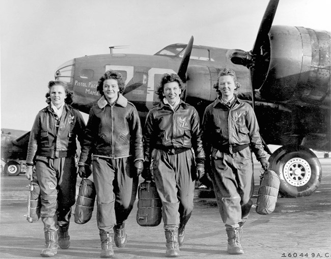 Four white women in leather jackets walk toward the camera away from a plane