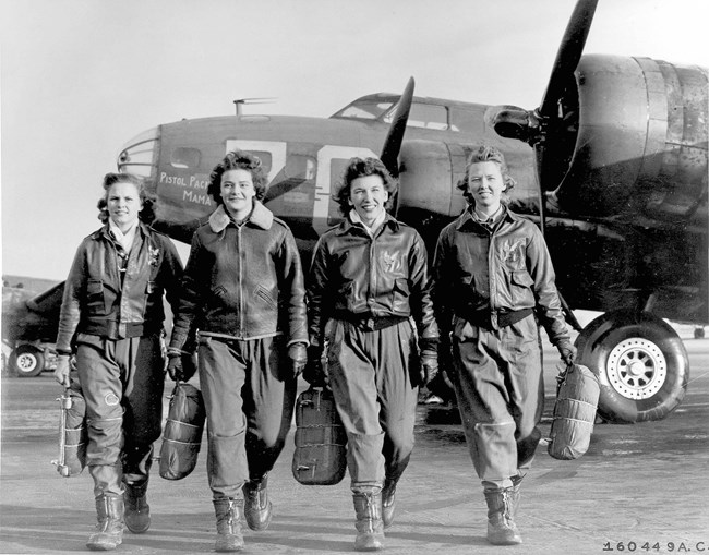 Four white women in leather jackets walk toward the camera away from a plane