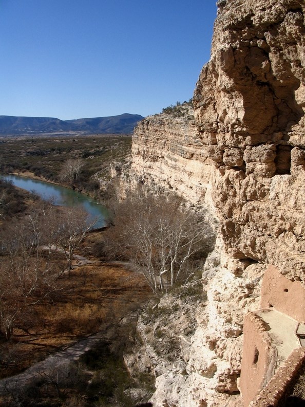 Guebard 2015 image View from Montezuma Castle, MONTEZUMA CASTLE NATIONAL MONUMENT. Red cliffs sweep from the right to the left distance, with green mountains in the background and a blue green river to the left.