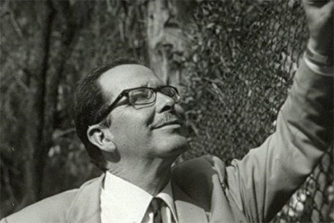 Guillermo Haro credit Elena Poniatowska slash Haciaelespacio.gob.mx Black and white photo of a man in glasses and a suit smiling as he looks up leaning against a fence