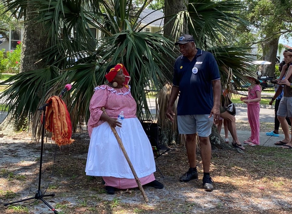 Large black woman in red gingham print dress with white apron and red headscarf sits holding a wooden walking stick.