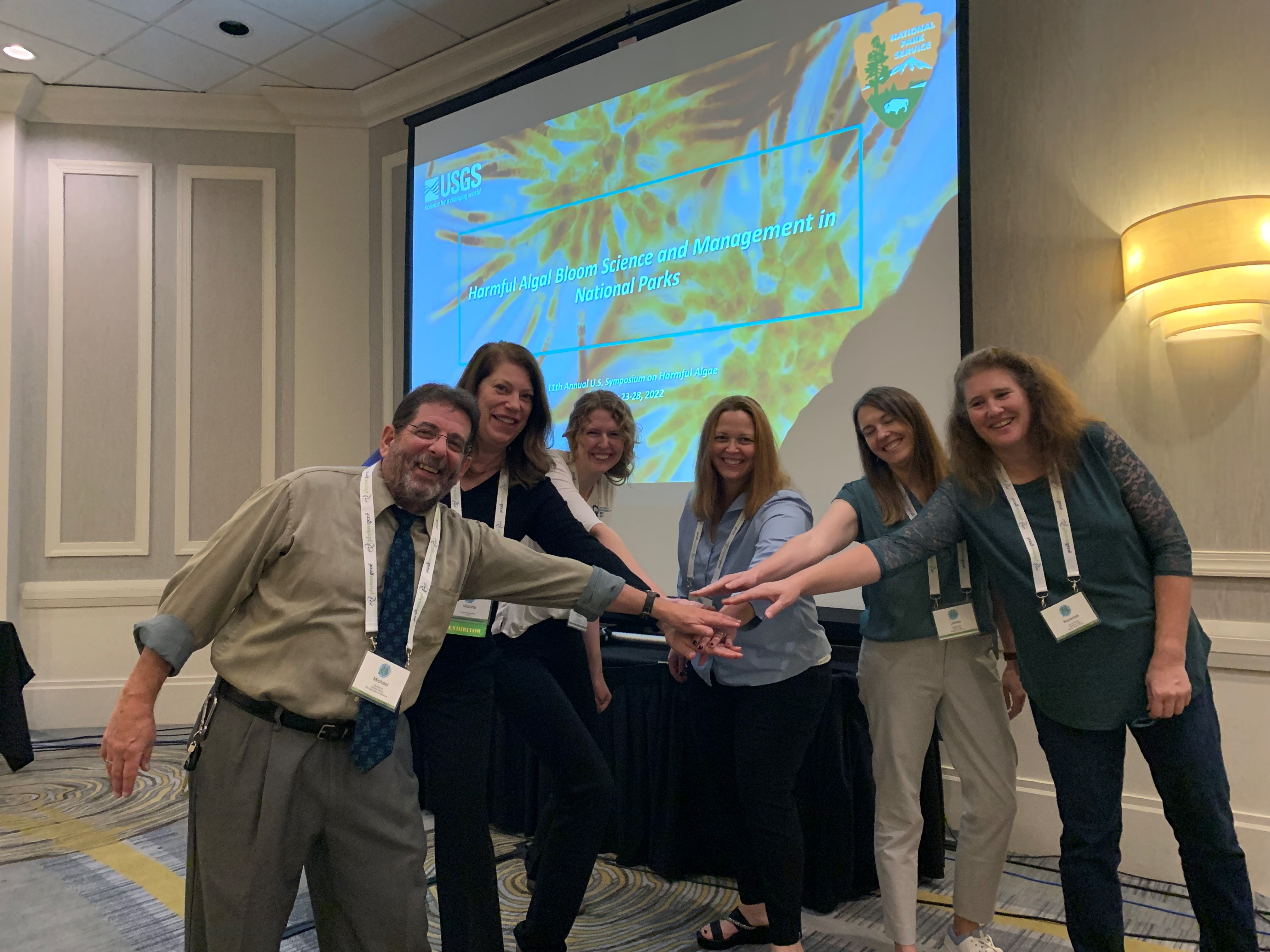 11th HABs symposium presentation a group of six presenters extend their arms and hands into the middle of their semi circle in a "go team" gesture in front of a projection screen with a title slide that says "harmful algal blooms science and management in national parks"
