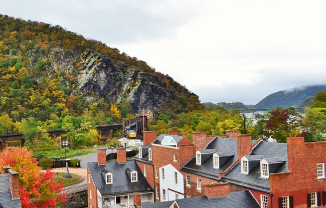 Brick buildings surrounded by trees in autumn, steep, rocky hill, river, train emerging from hill tunnel.