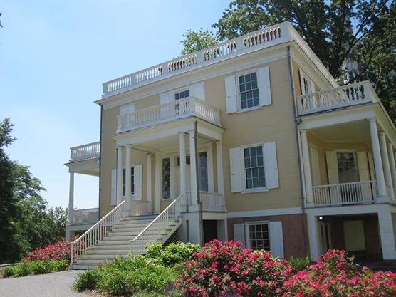 A large yellow house with white shutters sits in front of flowering bushes