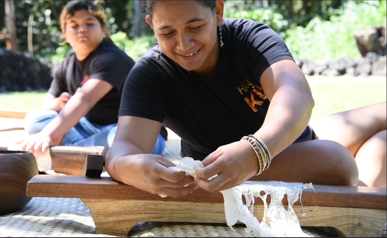 Still from a video of a woman bent over a wooden bench with a white cloth draped over it. She is sitting on a woven mat. She holds the cloth in place. Her other hand has a rectangular wooden instrument.