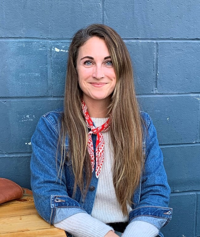 Woman with long brown hair sits in front of a blue cinderblock wall