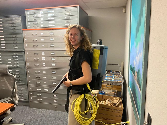 Woman wears backpack vacuum. Map cabinets are behind her.