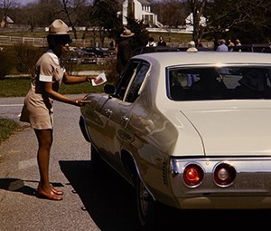 A women wearing a tan mini skirt dress with a Ranger hat is leaning towards a car window to hand something to the driver.
