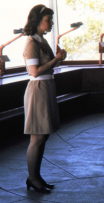A young woman wears a tan dress with a white color, white capped sleeves and a white belt and dark brown shoes.
