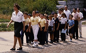 HFCA 1607_NPS Employees, Women_310_U_cropped A woman wearing a white blouse and dark green skirt leads a group of visitors on a walk. She is wearing oxford style shoes.