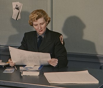 HFCA 1607_NPS Employees, Women_516_U_Cropped A woman wearing a NPS uniform sits at a desk reading a document. Her fram casts two shadows on the walls behind her.