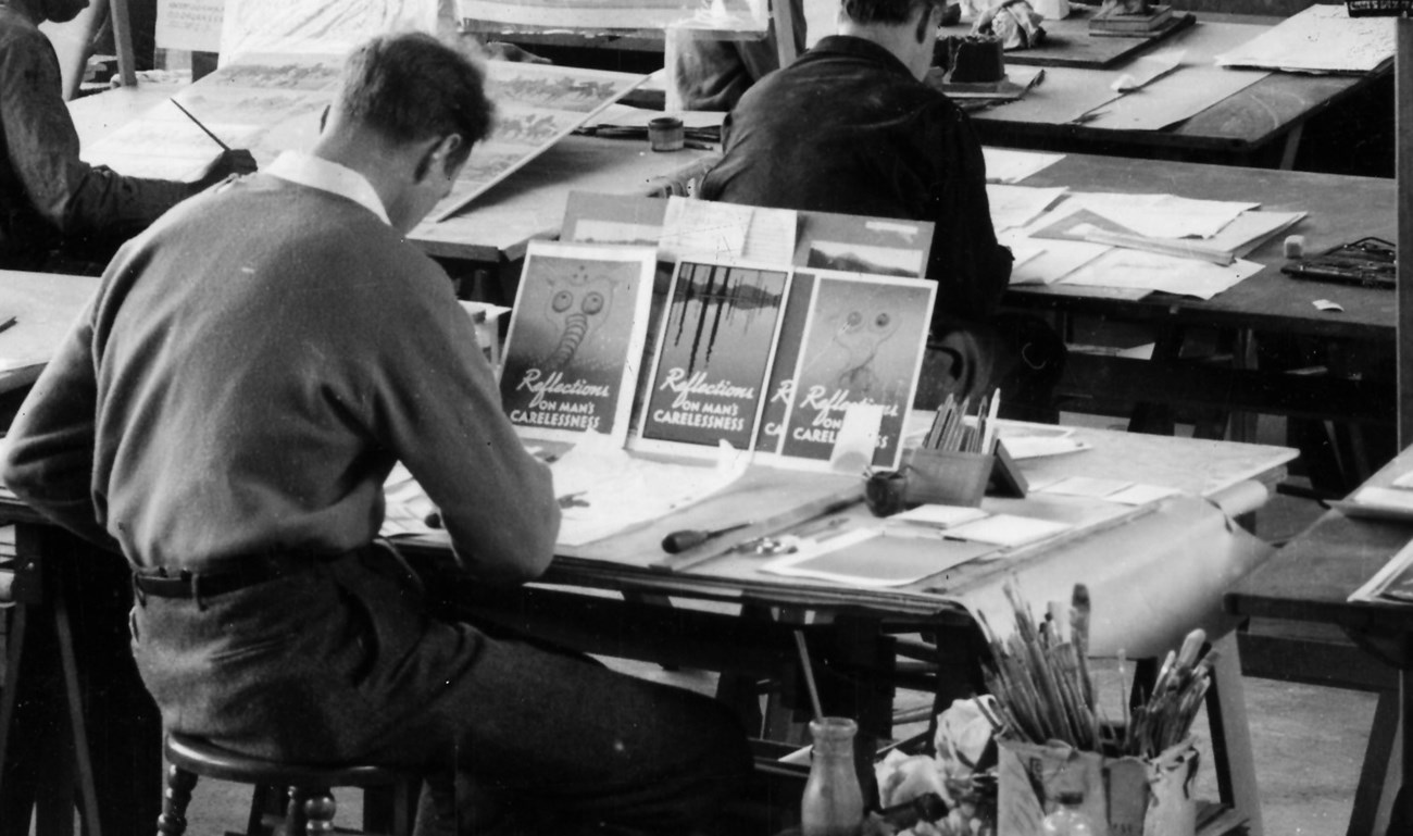 Man at desk (back turned) with 3 posters propped on desk