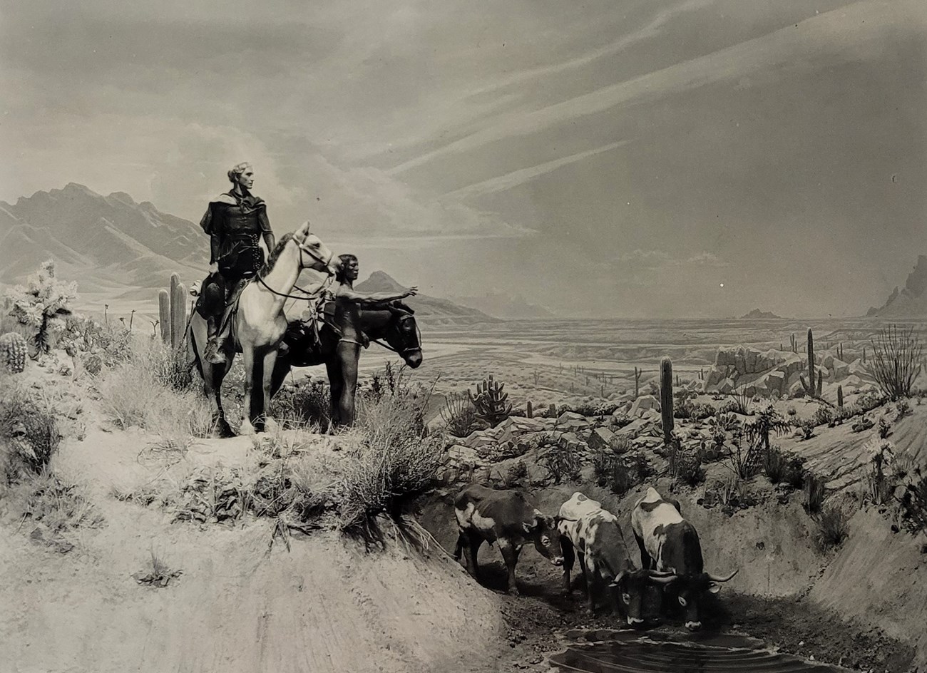 Desert scene diorama with two men on a horse on a hill above cows drinking from a stream below