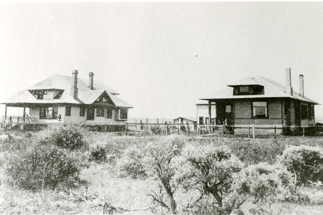 Black and white photo of two homes in sagebrush. The homes are side by side.