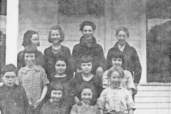 Twelve children gather on the steps of an old schoolhouse.