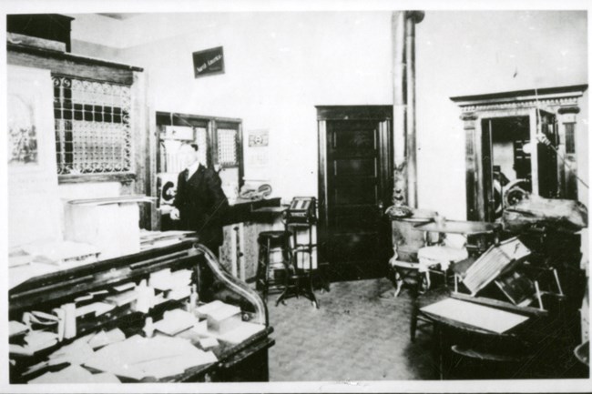 First National Bank of White Bluffs Black and white photo of the interior of a cluttered bank. A man in a suit stands at the center-left of the room.