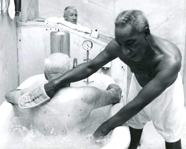 African American bath attendant scrubbing man's back in whirlpool tub in men's bath hall of Buckstaff Bathhouse; man in steam cabinet in background.