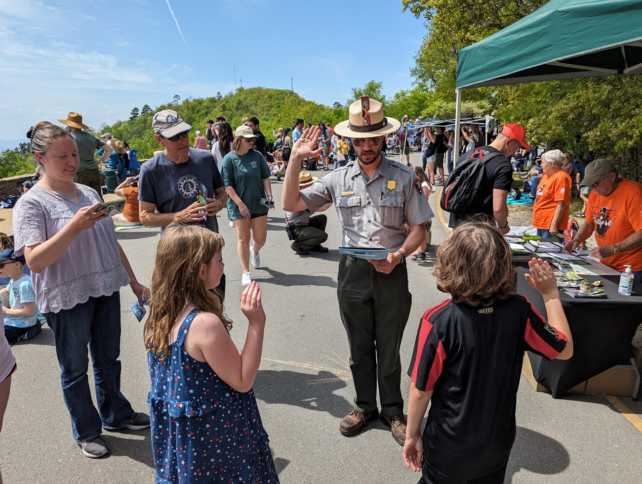 HOSP Junior Rangers Eclipse Park ranger in uniform raises right hand and leads two kids in Junior Ranger pledge. Their family looks one with smiles. Behind is an eclipse event table set up and crowd.