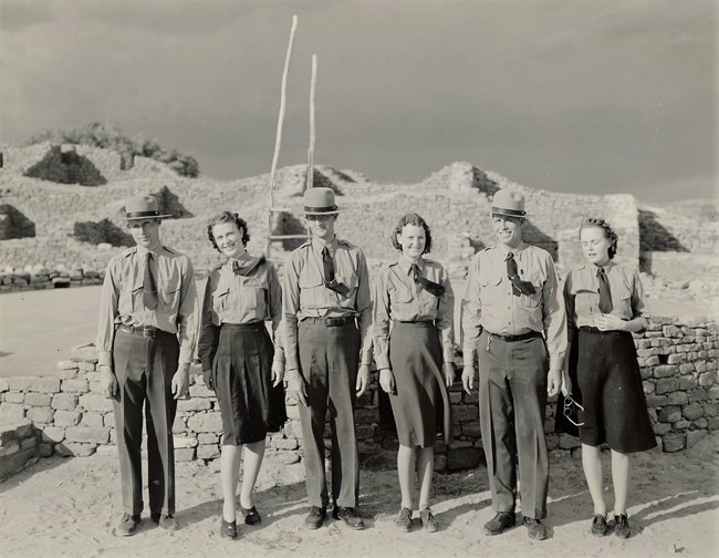 Three men and three women in NPS uniforms stand in a line, looking towards the camera. They pose in front of an underground stone structure with a tall wooden ladder coming out of it.