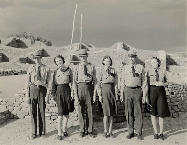 Aztec Ruins staff Three men and three women in NPS uniforms stand in a line, looking towards the camera. They pose in front of an underground stone structure with a tall wooden ladder coming out of it.