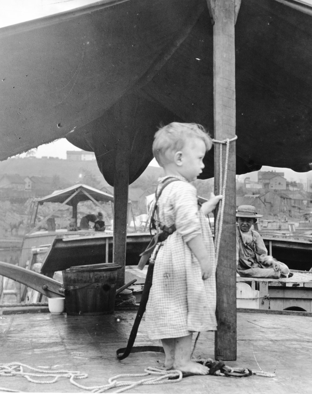 Small child stands on the deck of a boat. A rope ties him to a mast.
