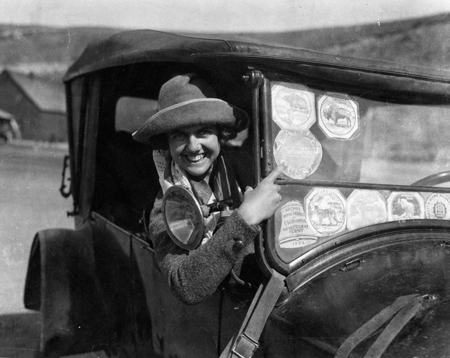 Woman in an old car points to stickers on the windshield