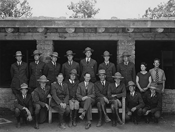 A group of men and one woman pose in two rows.  The first row is all men and some are kneeling and some are seating on a bench. The second row is positioned just behind them.  the group is in front of a building.