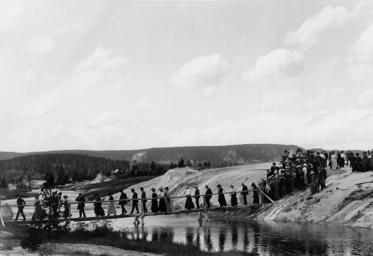 Large group of men and women cross a log bridge