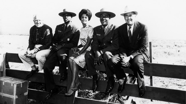 Cast of Spenser's Mountain Four men and a woman sitting on a fence. The men include two park rangers in uniform and actor Henry Fonda.