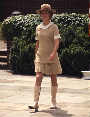 A woman wearing a tan dress, tan hat and tan knee boots walks towards us.