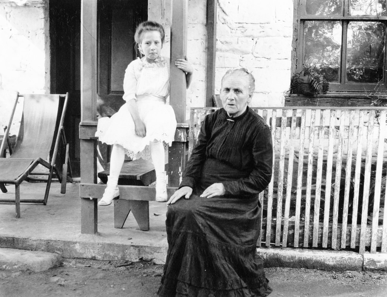 Old woman and young girl sit in front of a house