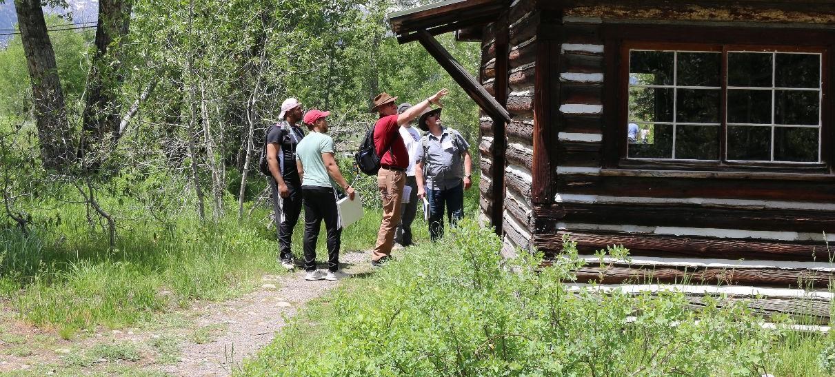 A group of five people stand on a dirt path, leading through low vegetation to a one-story log cabin