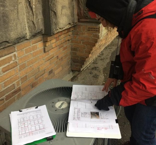 A person looks at drawings in a binder and clipboard on an air-conditioning unit, beside a brick wall.