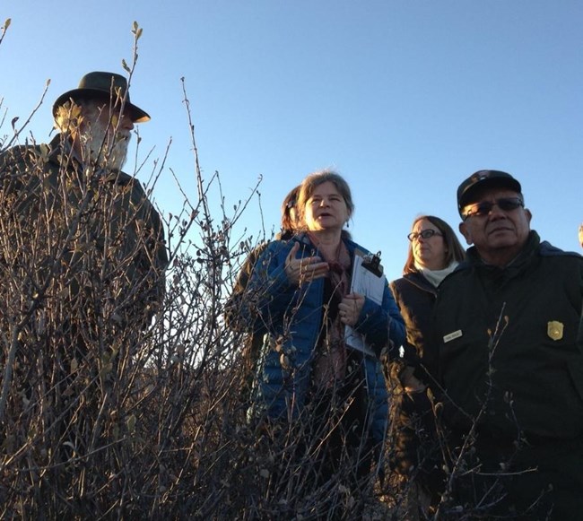 A group of people, some in NPS uniform, stand in a field behind dry vegetation under clear blue sky. The person in the center speaks and holds a clipboard.