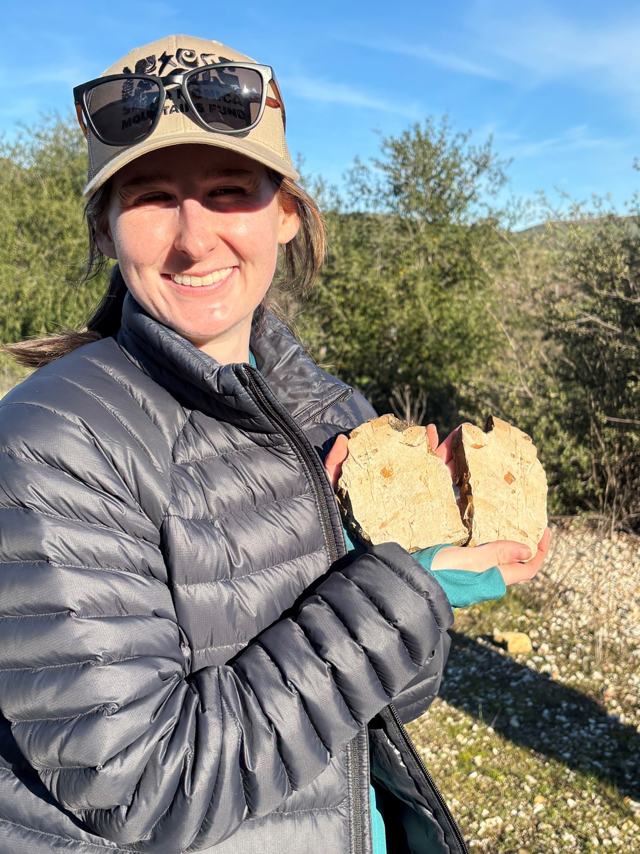 A smiling person standing outdoors holds two hand-sized pieces of rock that have split from each other. Fossil bones and fish scales are present on the rock surfaces.