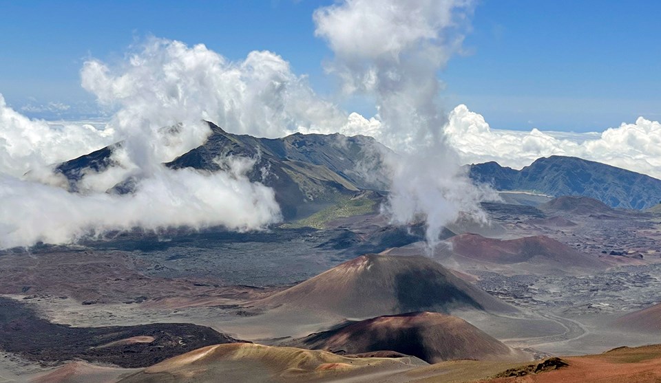 A close-up of the inside of the crater with a red and brown hill in the bottom right of the image, multi-colored cinder cones in the middle, and dark green mountainous areas in the background.