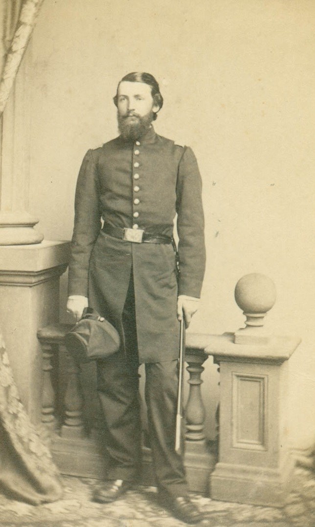 bearder man in uniform standing holding his cap and sword.