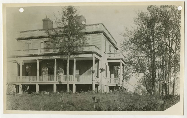 A historic black and white photo of a house next to a cluster of bare trees to its right.