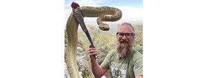 Bryan with a beard, glasses, and t-shirt, using a metal tool to hold up an enormous snake.