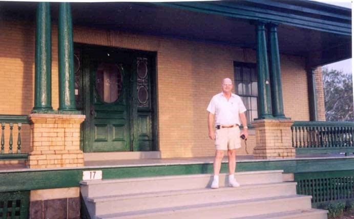 an older man in white shirt and short stands on the porch of a historic building