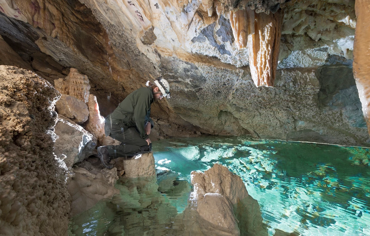 Hansen Cave pool A uniformed National Park Service employee with a helmet and light kneels on a cave floor overlooking a cave pool