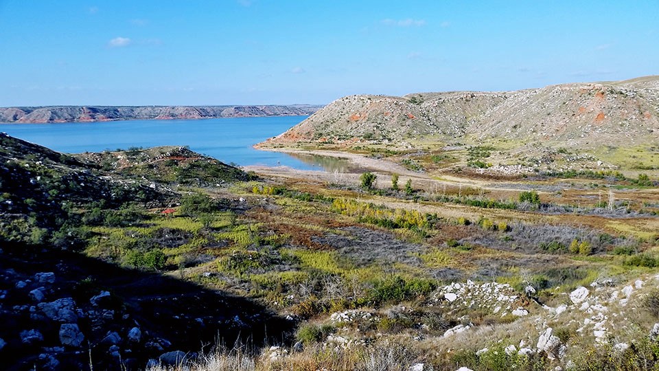 View from a hill of a valley of low, scrubby, desert plants, barren hills, and a bright blue lake.