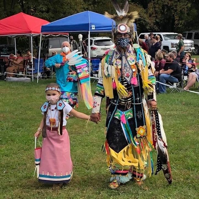 A man and a young girl holding hands and wearing traditional Native regalia.