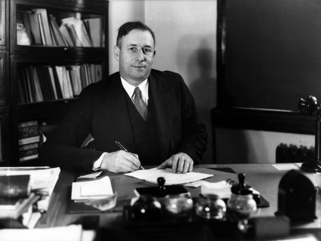 Harold Bryant Man in suit sitting at a desk looks up towards camera